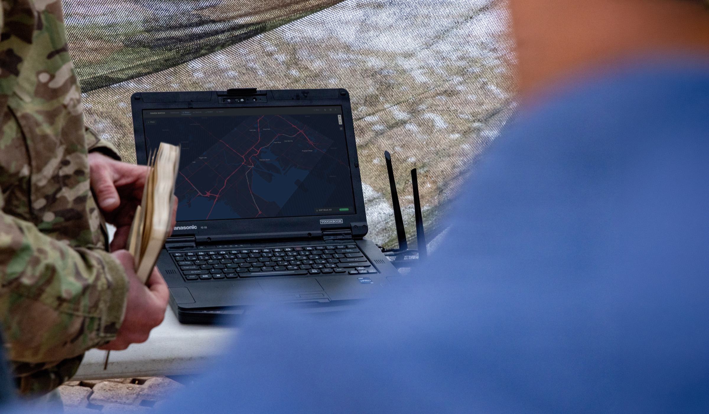 An operator working a ruggedized laptop under camouflage netting